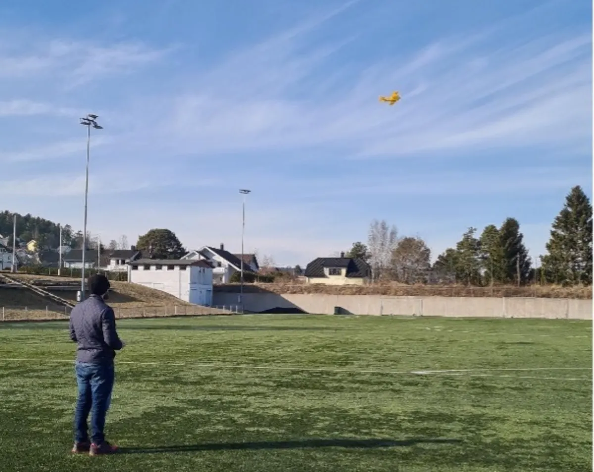 Man flying a small yellow drone on a sports field with houses in the background