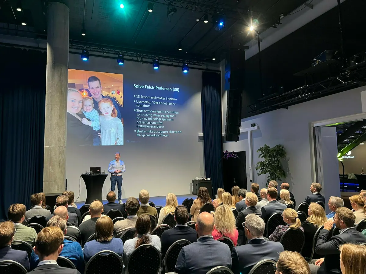 Presenter on stage beside a slide with a man's photo and his bio, audience seated