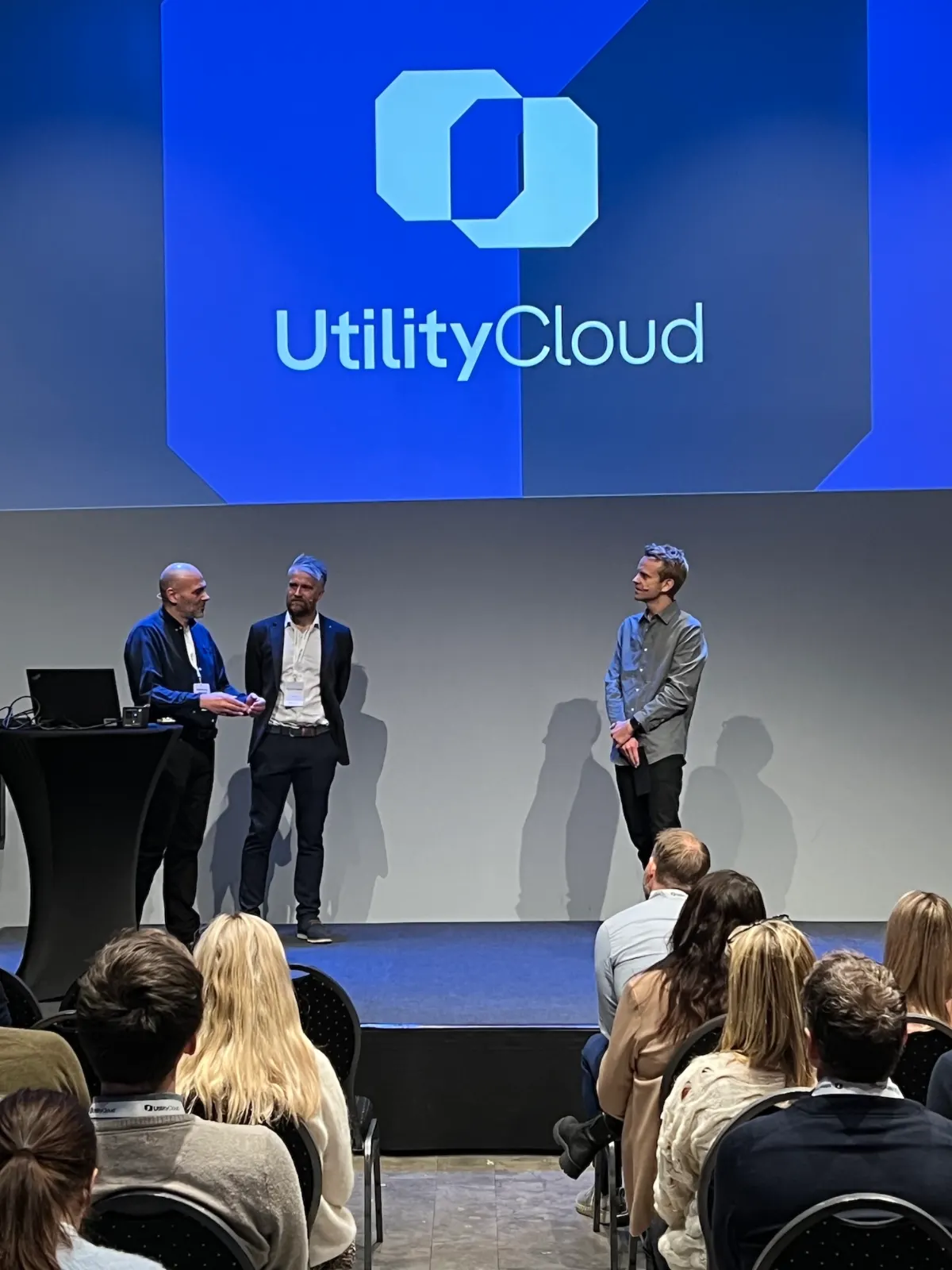 Three men on stage under a large UtilityCloud logo, one speaking to another