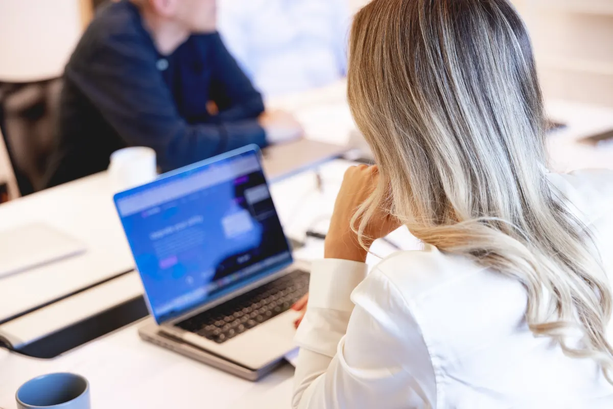 Woman seen from behind working on a laptop at a meeting table with colleagues