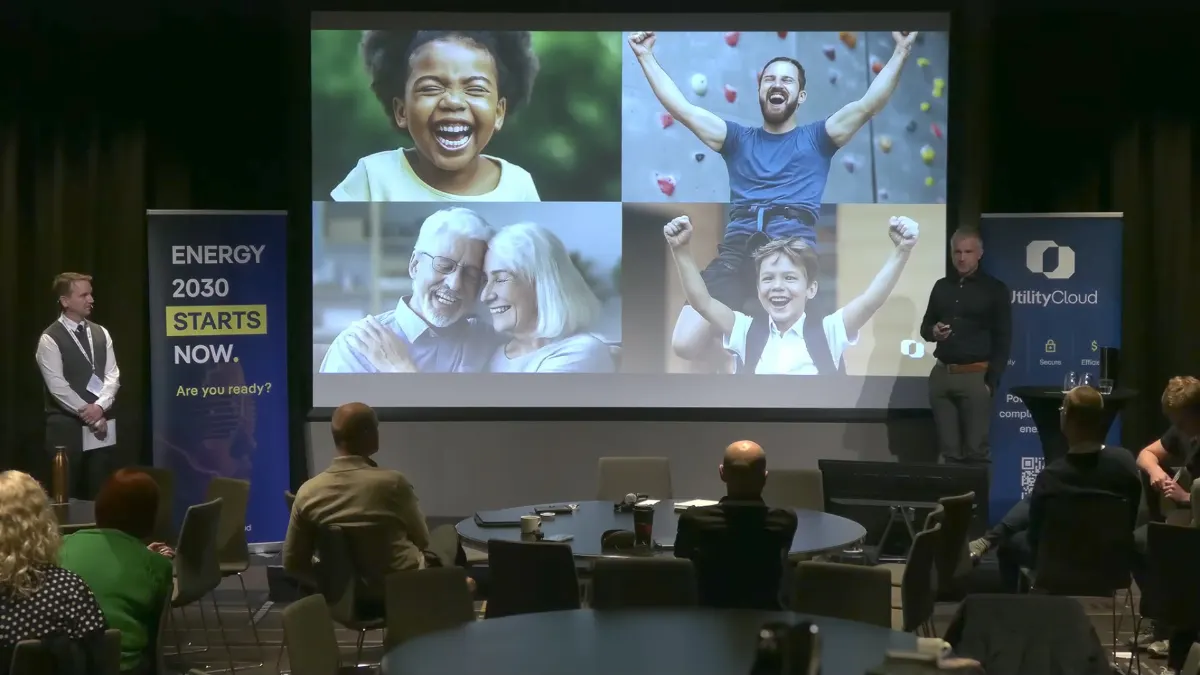 Conference stage with four smiling people on screen and audience at round tables